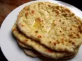 Stos świeżo upieczonych, lekko przypieczonych placków roti i naan, polanych roztopionym masłem, na białym talerzu.