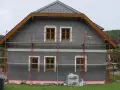 Dom w trakcie ocieplania. Szara elewacja, drewniane okna, rusztowanie. Materiały budowlane Austrotherm wskazują na dbałość o niski współczynnik przewodzenia ciepła.