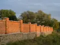 Solidny płot murowany z cegły i kamienia, z ozdobnymi słupkami, otacza teren porośnięty zielenią.