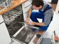 Fachowiec w niebieskim kombinezonie montuje element zmywarki, pokazując, jak podłączyć zmywarkę. Obok leżą klucze i rękawice.