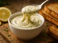 Une cuillère de sauce au comté crémeuse et fumante, parsemée d'herbes, est retirée d'un bol blanc. Des toasts grillés et des citrons sont en arrière-plan.