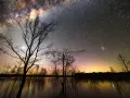 Canon EOS 2000D photo de nuit : une voie lactée spectaculaire au-dessus d'un lac reflétant les arbres dénudés et un ciel orangé.