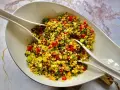 Kolorowa sałatka z kaszą bulgur, pomidorami, ogórkiem i ziołami, podana w białej misie z dwoma białymi szczypcami.