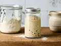 Blubbernder Sauerteig im Glas, bereit zum Backen. Daneben ein Glas mit Mehl, das zeigt, wie man den Sauerteig pflegen kann.