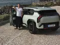 Mężczyzna opiera się o drzwi nowego, jasnoszarego Kia EV3, zaparkowanego na brukowanej drodze nad morzem.