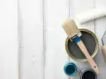 Herramientas para pintar mesitas de noche: lata de pintura gris, pincel, rodillo y botes de pintura azul y negra sobre fondo de madera blanca.