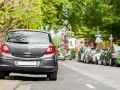 Graues Auto parkt am abgesenkten Bordstein. Mehrere Autos reihen sich die Straße entlang unter grünen Bäumen.