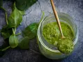 Słoik z zielonym pesto, na łyżce porcja sosu. Czy pesto ze słoika się podgrzewa? Świeże liście bazylii obok.