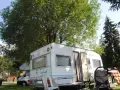 Przyczepa kempingowa na polu namiotowym Jonidło, otoczona zielenią. Obok wózek i leżak.