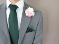 Elegancki pan w szarym garniturze, białej koszuli, zielonym krawacie i poszetce. Na klapie marynarki ozdobna brustasza z różowych i białych kwiatów.