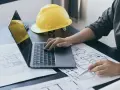 Architekt pracuje nad projektem, analizując plany i obliczając powierzchnię całkowitą budynku. Żółty kask ochronny leży obok.