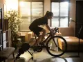 A woman enjoys indoor cycling workouts at home, following a workout on her tablet. Sunlight streams through the window, illuminating her focused effort.