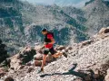 Un corredor de trail desciende por un sendero rocoso y empinado, con montañas boscosas al fondo.