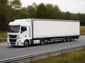 Biały tir z naczepą jedzie po drodze, otoczony zielenią.
