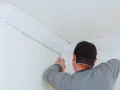 Mężczyzna w czapce montuje białą listwę styropianową w rogu pokoju.