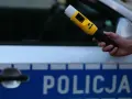 Dłoń trzymająca żółty alkomat na tle radiowozu policyjnego z napisem "POLICJA".