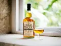 Ein besonderer Rum, "The Real McCoy", steht neben einem Glas mit bernsteinfarbener Flüssigkeit vor einem Fenster mit grünem Bokeh.