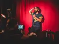 Ein Comedian steht auf einer Bühne in Berlin, hält ein Mikrofon und wischt sich die Stirn. Ein Schild mit "Chips und Kaviar" steht daneben.