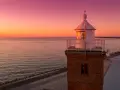 Zachód słońca nad morzem w Darłówku Wschodnim. Latarnia morska oświetla spokojne wody, a na promenadzie spacerują ludzie.