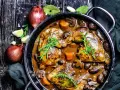 Herzhaftes Coq au Vin in einem Gusseisentopf, mit Pilzen, Karotten und Kräutern garniert. Daneben Zwiebeln und Knoblauch.