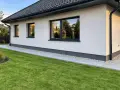 Nowy dom z białym tynkiem i czarnym dachem. Wokół domu ścieżka z płyt i zielona trawa. To najtańsza opaska wokół domu, która dodaje uroku.