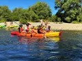Des personnes profitent d'une activité canoe kayak sur une rivière ensoleillée, entourées de verdure et d'une plage de galets.