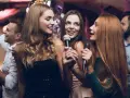 Trzy kobiety śpiewają do mikrofonów, bawiąc się przy piosenkach do śpiewania. W tle widać innych ludzi na imprezie.