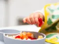 Rączka 9 miesięcznego dziecka sięga po kawałek truskawki z miseczki pełnej owoców.