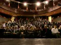 Widownia teatru, ludzie siedzą na miejscach, widać balkon z widzami.