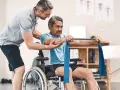 Ein Mann im Rollstuhl trainiert mit einem Therapeuten, der ihm hilft, die Widerstandsbandübungen auszuführen. Die Physiotherapeuten unterstützen ihn bei seiner Genesung.