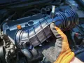 Gloved hand holds a black intake hose near a car engine. This part is often replaced during vacuum leak repair, affecting the vacuum leak repair cost.