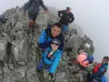 Grupa turystów na skalistym szczycie, otoczona mgłą. Jedna z kobiet, **mała wysoka**, robi sobie selfie z partnerem.