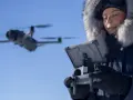 Woman in a fur-lined hood controls a flying drone in cold weather, looking at a tablet.