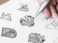 Main d'un designer esquissant des idées pour comment dessiner un logo. Plusieurs croquis abstraits sont visibles sur le papier.
