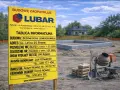 Żółta tablica informacyjna budowy z danymi o inwestycji i logo firmy LUBAR na tle placu budowy.