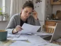 Kobieta zamyślona przegląda dokumenty, szukając wzoru wniosku o upadłość konsumencką. Na stole laptop i kubek kawy.
