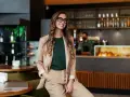Mujer sonriente con gafas, vestida de traje beige, sentada en un restaurante moderno. Detrás, un barista trabaja. Esencial para la gestión de restaurantes.