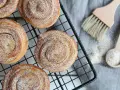 Nybakade karlsbaderbullar med socker och kanel på ett svart galler. En borste och en sked med socker ligger bredvid.