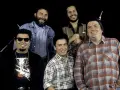 Los Lobos, native sons, pose together. One man wears sunglasses and a fringed jacket, another a denim jacket, and two sport plaid shirts. A saxophone is visible.