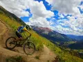 A mountain biker navigates a steep, winding trail, experiencing the best mountain biking in Colorado with stunning valley views and dramatic clouds.