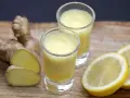 Zwei Gläser mit leuchtend gelbem Ingwer Shot, daneben frischer Ingwer und Zitronenscheiben auf Holzbrett.