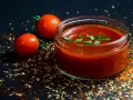 Un vasetto di ketchup fatto in casa, guarnito con basilico fresco, accanto a pomodori maturi e spezie su sfondo scuro.