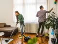 Pareja colaborando para purificar casa. Ella aspira el suelo, él quita el polvo de las paredes.