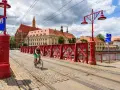 Mężczyzna na rowerze jedzie po brukowanej drodze rowerowej z torami tramwajowymi, w tle zabytkowe budynki i czerwony most.