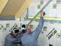Dwóch budowlańców pracuje przy konstrukcji szkieletowej. Jeden mierzy taśmą, drugi zaznacza.