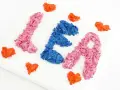 Comment faire du relief sur un tableau : des lettres "LÉA" en papier froissé rose et bleu, entourées de cœurs orange sur une toile blanche.