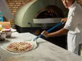 Pizza cruda en pala, lista para horno de leña con chef al fondo.