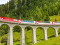 A red and white train travels across a stone viaduct, winding through a lush green valley with a dense forest on the mountainside. This scenic route is part of the Amsterdam to Basel train journey.