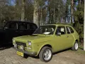Zielony fiat 127 z lat 70. stoi obok czarnej taksówki na tle drzew.