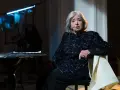 Stara kobieta w błyszczącej sukience siedzi na krześle, opierając się o stół z nutami.